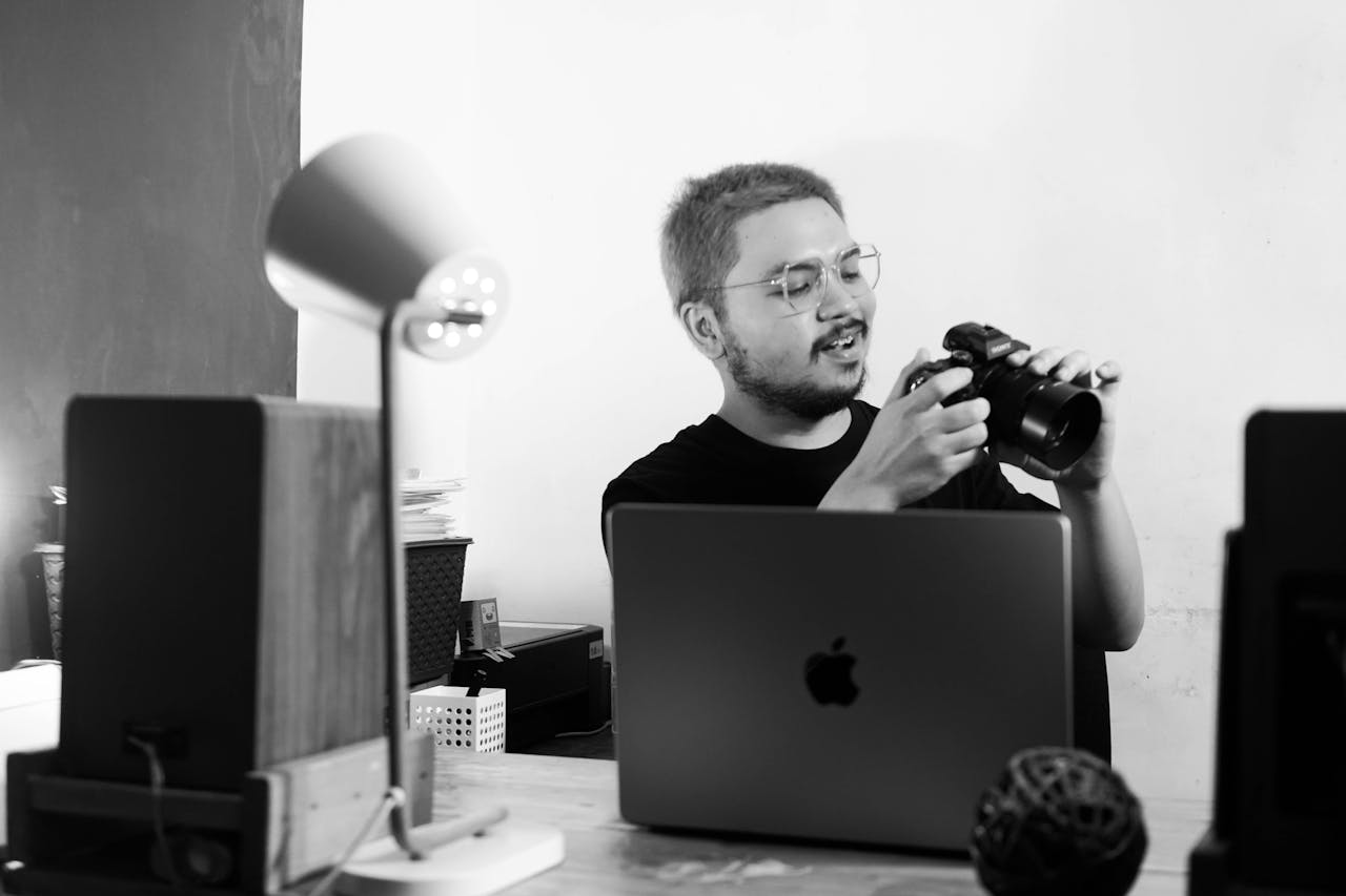 Home Black and white photo of a man reviewing a camera at his desk in a modern office environment.