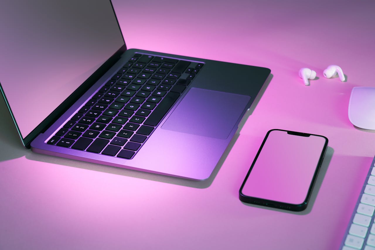 Stylish tech workspace featuring a laptop, smartphone, and earphones under pink lighting.
